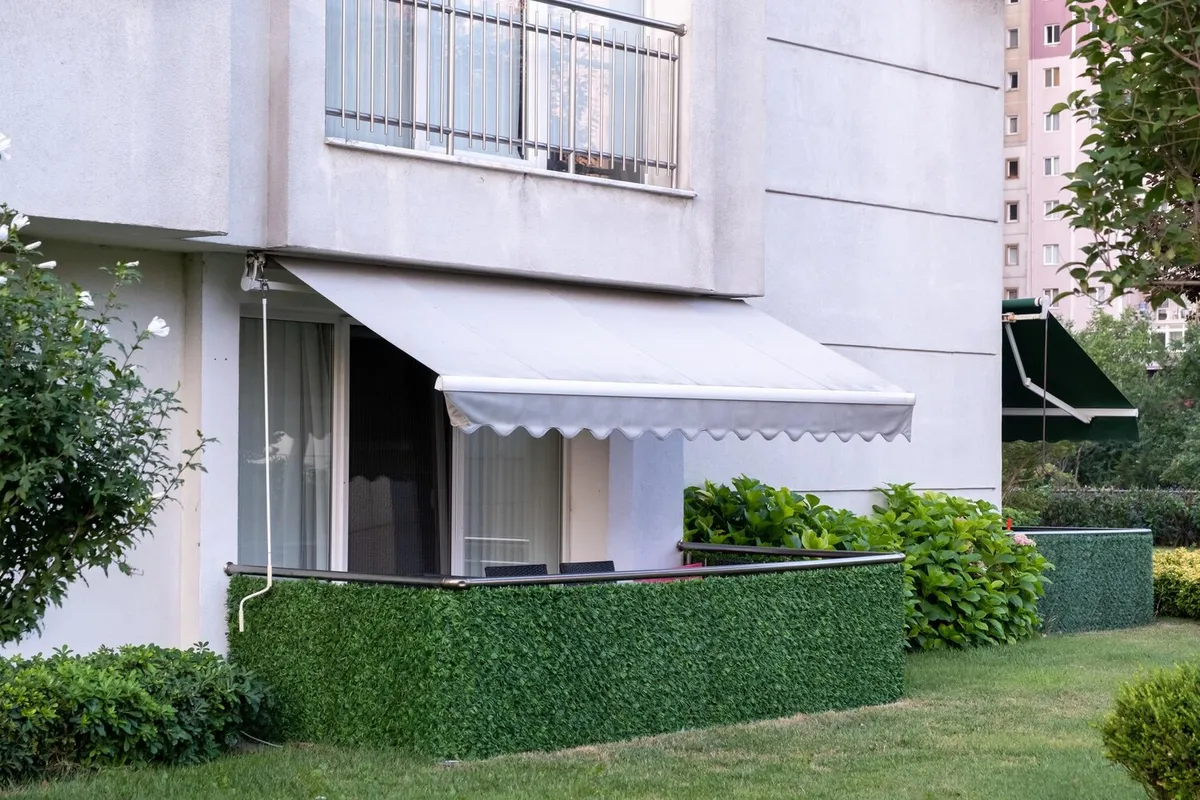 Store banne à toile gris clair déployé au-dessus de la terrasse en rez-de-jardin d'un immeuble, un second store vert en arrière-plan